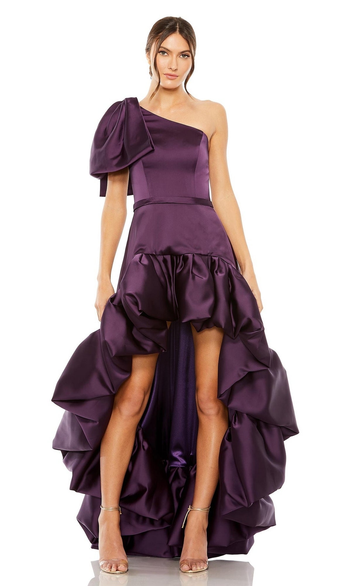 A woman wears the High-Low Party Dress 20702 by Mac Duggal, featuring an aubergine purple one-shoulder design, high-low ruffled skirt, large side bow, satin finish, and pairs it elegantly with nude heels.